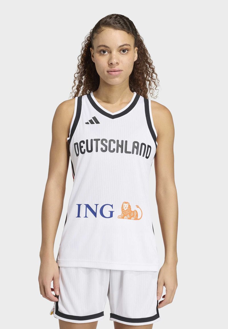 Jeune femme debout, portant un maillot de basketball blanc de l'Allemagne avec une bordure noire et le logo ING à l'avant, ainsi que des shorts assortis.