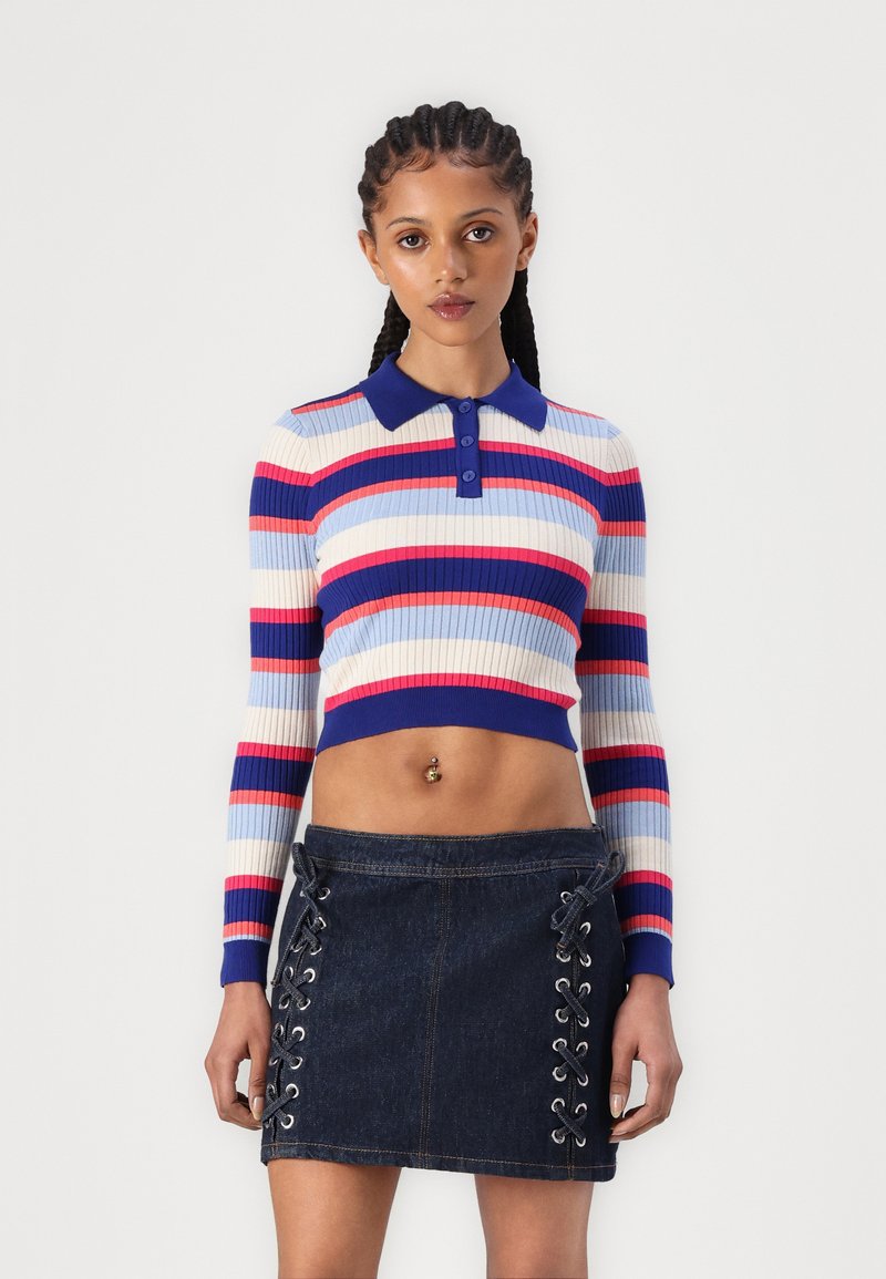 Pull cropped rayé avec des couleurs bleue, rouge et rose ; dispose d'un col et d'une patte de boutonnage. Jupe en denim foncé avec des détails à laçage sur les côtés.
