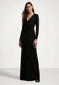 Svart langermet kjole med dyp V-hals og knutedetalj i midjen. Stoffet er glatt og tettsittende, noe som fremhever silhuetten.