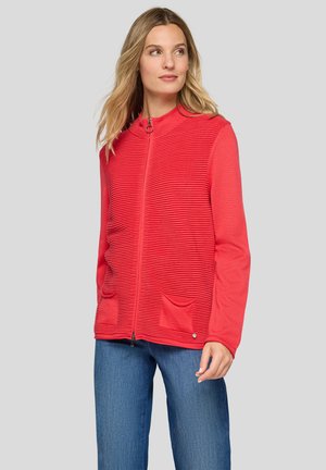 Frau trägt eine rote, texturierte Jacke mit Reißverschluss und Taschen sowie blaue Jeans, blickt seitlich vor einem schlichten hellgrauen Hintergrund.