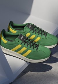 Pair of green Adidas sneakers with yellow stripes and black laces resting on a white geometric surface in soft light.