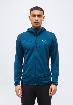 Muž v modrej bundoškrupinovej bunde s kapucňou, s oranžovými sťahovacími zipsami a bielym logom na hrudi, stojaci pred jednoduchým pozadím.