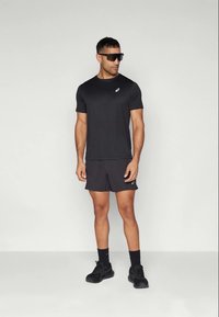Schwarzes sportliches T-Shirt und Shorts, strukturierten Stoff, figurbetontes Design, reflektierendes Logo auf dem Shirt, kombiniert mit schwarzen Turnschuhen und Sonnenbrille.