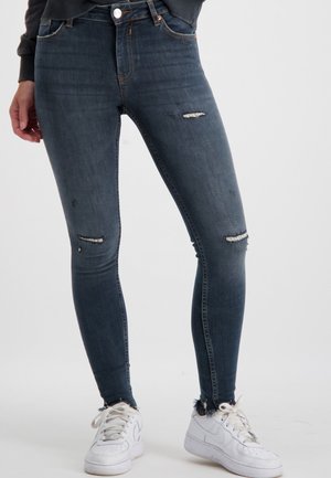 Dunne donkerblauwe denim jeans met gescheurde details op de knieën, versleten zomen en een klassiek vijf-pocket ontwerp. Gepaard met witte sneakers.