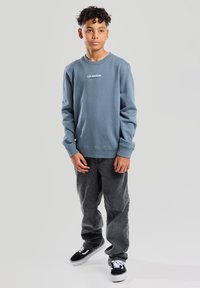 Sudadera azul de cuello redondo con el texto "LOS ÁNGELES", combinada con pantalones gris oscuro y zapatillas negras. Tela suave, corte relajado, mangas largas.