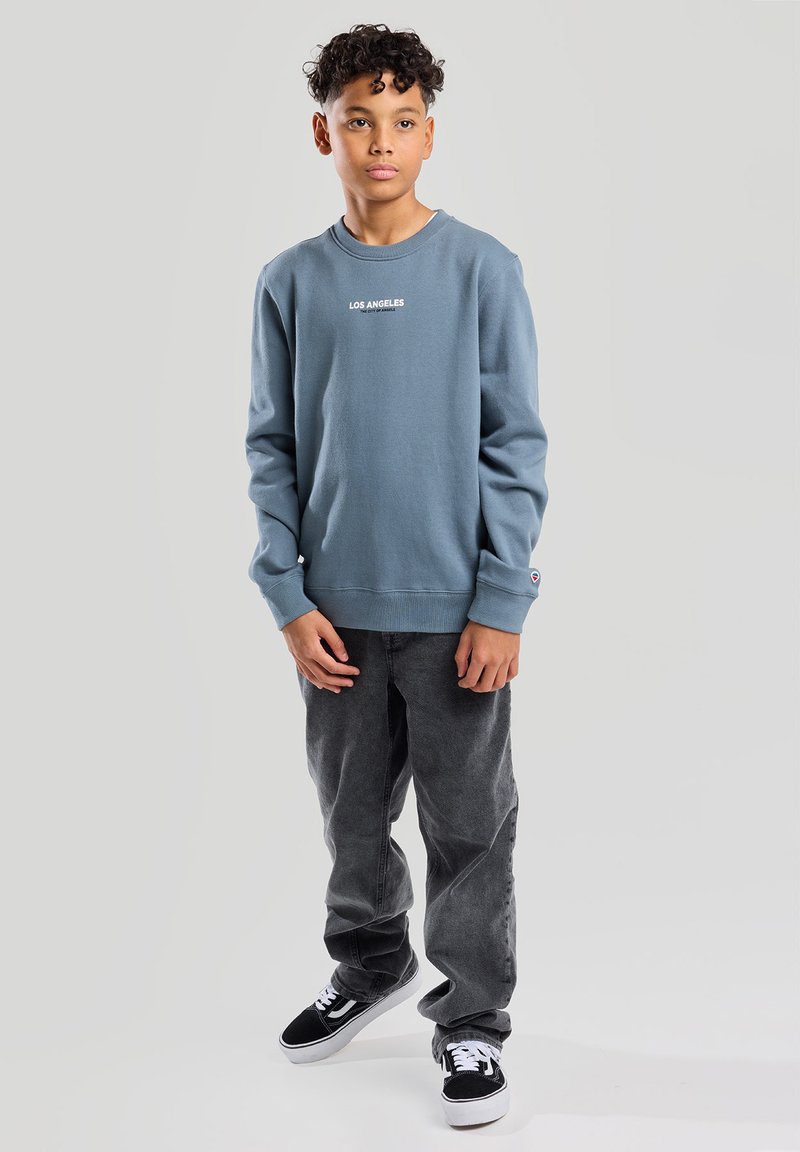 Sudadera azul de cuello redondo con el texto "LOS ÁNGELES", combinada con pantalones gris oscuro y zapatillas negras. Tela suave, corte relajado, mangas largas.