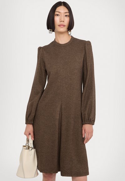 Ruskea herringbone-mekko, jossa on pitkät puffihihat, korkea kaulus ja laskostettu etuosa. Mukana vaaleanbeige käsilaukku, jossa on hopeiset yksityiskohdat.