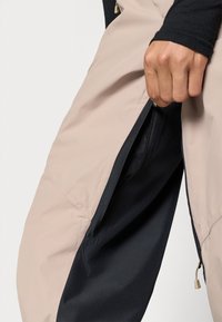 Pantaloni bej deschis cu garnitură neagră, având un fermoar pe lateral. Materialul este neted și are un ușor luciu, potrivit pentru activități în aer liber.