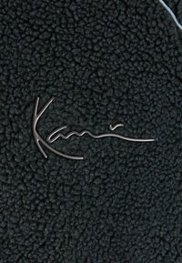 Black textured fleece fabric with a prominent silver metallic logo, featuring smooth and curved lines in an organic design.
