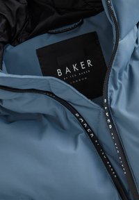 Mėlynos striukės vidus, rodantis juodą etiketę su užrašu "BAKER by Ted Baker London" ir dalinį juodo pamušalo bei užtrauktuko vaizdą.
