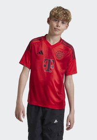 Maillot de soccer rouge à manches courtes, avec des accents noirs, un col en V et un motif texturé. Affiche le logo et des éléments graphiques de manière proéminente.