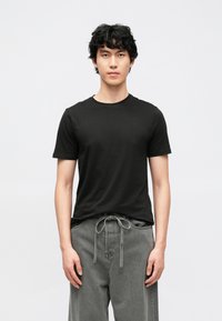 Črna bombažna majica s kratkimi rokavi in okroglim vratnim izrezom, kombinirana z sivimi hlačami z vezalko, stoji pred preprostim ozadjem.