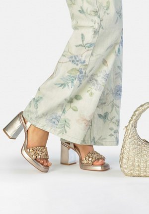 Pies con pantalones anchos de flores que llevan sandalias doradas de tacón ancho con tiras de gemas junto a un pequeño bolso dorado texturizado.