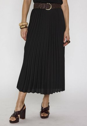 Jupe midi plissée noire avec une ceinture tissée marron, sandales marron à talons hauts et bracelets dorés au poignet gauche.