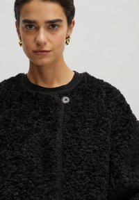 Zwarte textuurjack met een fluffy materiaal, ronde halslijn en knoopsluiting. Bevat een gebreide rand rond de halslijn en voorkant.