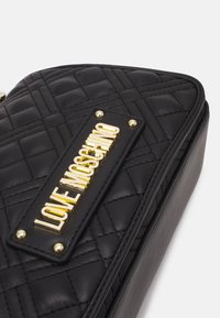 Svart vadderad läderväska med guld "LOVE MOSCHINO" logoplack och dekorativ sömnad mot en ren vit bakgrund.
