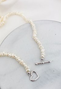 Witte parelketting met onregelmatige vormen, met zilveren hart- en toggle-sluiting accenten, op een gladde, ronde ondergrond.