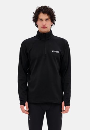 Junger Mann mit lockigem Haar, der ein schwarzes Langarm-Quarter-Zip-Oberteil und schwarze Leggings mit weißem cep-Logo trägt.