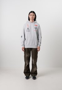 Hanorac gri cu glugă, decorat cu logo-uri și text, asortat cu pantaloni cargo negri, prevăzuți cu buzunare. Pantofii sport negri completează ținuta.
