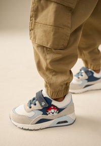 Sneakers med en blanding af blå, grå og hvide materialer; har en velcrolukning og et karakter-patch på siden. Tekstureret stof og polstret design.