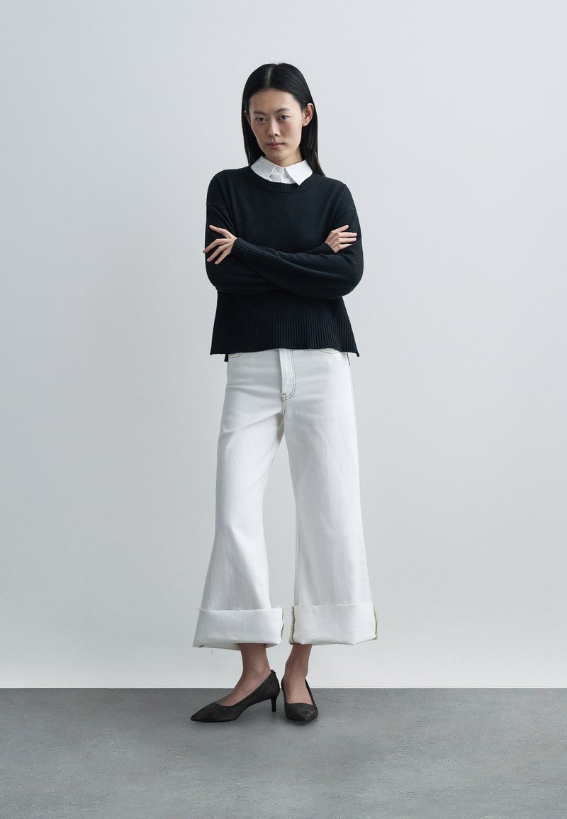 Pull noir en maille avec texture côtelée et encolure ronde, associé à un jean blanc large et court avec ourlet effiloché. Talons noirs.