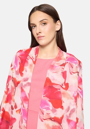 Young woman with long brown hair wearing a pink dress and a vibrant floral shawl in red, pink, and white tones.