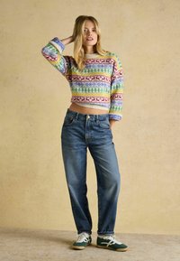 Gestrickter, kurz geschnittene Pullover mit buntem Muster, kombiniert mit hoch-taillierten blauen Jeans. Das Modell trägt grüne Sneakers mit weißen Akzenten.