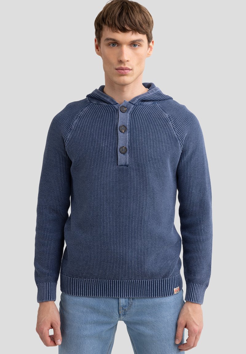 Tamsiai mėlyna gobtuvinė džemperis su tekstūruotu mezgimo raštu, trys mygtukai prie kaklo, ir raštuoti rankogaliai bei apačia. Dėvimas ant šviesiai mėlynų džinsų.