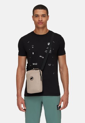 Beżowa torba crossbody z logo, zamykana na zamek, wykonana z trwałego materiału, z regulowanym paskiem, noszona przez osobę w czarnej koszuli.