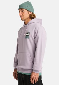 Felpa con cappuccio lavanda, dotata di tasca frontale e logo ricamato. Indossata con una maglietta verde menta sotto e un berretto di maglia turchese chiaro.