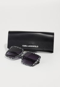 Black sunglasses with a square shape, featuring a white leopard print frame and dark lenses, accompanied by a black case with "KARL LAGERFELD" logo.