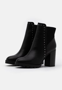 Botines negros con tacón grueso, material de cuero liso, parte superior de tela acanalada y acentos con tachuelas en la parte delantera. Suela texturizada.