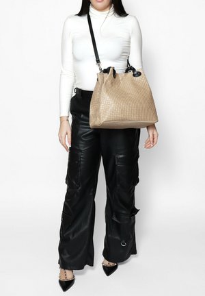 Geweven beige handtas met textuurpatroon, zwarte accenten en een enkele schouderband. Model draagt zwarte leren broek en een witte top.
