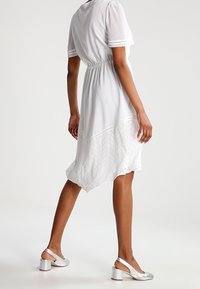 Valkoinen, lyhythihainen mekko, jossa istuva yläosa ja virtaava, epäsymmetrinen helma, jossa pitsikoristeita. Yhdistetty hopeisiin lohkokorkoisiin kenkiin.