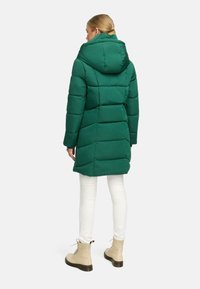 Giacca a vento verde scuro con cappuccio, texture trapuntata, design di lunghezza media, abbinata a pantaloni bianchi e stivali con lacci beige.