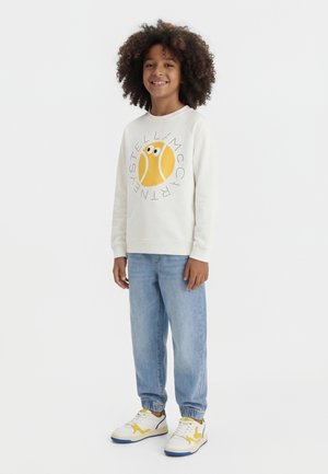 Barn med krøllet hår iført hvid sweatshirt med gul tennisboldgrafik, lyseblå jeans og hvide sneakers med gule detaljer.