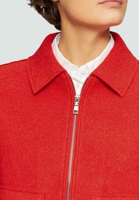 Czerwona kurtka z wełny z dodatkiem innych włókien o teksturowanym wykończeniu, z charakterystycznym kołnierzem i srebrnym metalowym zamkiem. Brak widocznych wzorów lub akcentów.