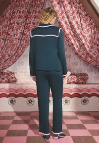 Mörkgrönt pyjamaset med rosa volangkrage och manschetter. Modell visad bakifrån, med blomsterprydda gardiner och rosa rutig golv i bakgrunden.