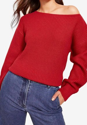 Femme portant un pull côtelé rouge épaules dénudées et un jean bleu taille haute, une main glissée dans la poche.