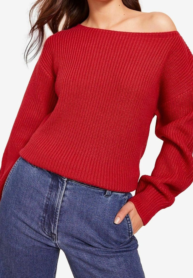 Femme portant un pull côtelé rouge épaules dénudées et un jean bleu taille haute, une main glissée dans la poche.