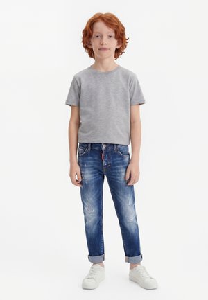 Jeune garçon aux cheveux roux se tenant face à l'objectif, vêtu d'un t-shirt gris, d'un jean bleu retroussé et de baskets blanches sur fond blanc uni.