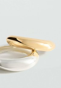 Dva glatka, zaobljena metalna prstena, jedan zlatni i jedan srebrni, koji se preklapaju na svijetloj neutralnoj pozadini.