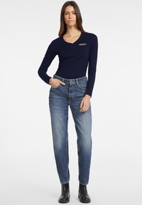 Navy lange mouw top met een V-hals en klein logo, gecombineerd met lichtblauwe rechte jeans en zwarte enkellaarsjes.