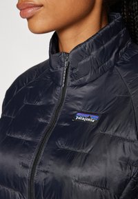 Veste matelassée synthétique bleu marine avec un col haut et une fermeture éclair, présentant des coutures horizontales et un logo Patagonia sur la poitrine.