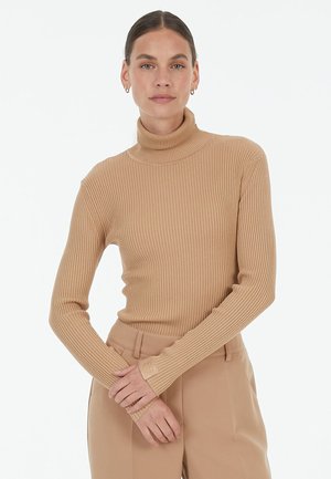 Beige ribbed turtleneck sweater with fitted long sleeves and a textured finish. Features a small logo on the cuff, paired with beige trousers.