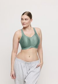 Mujer con un sujetador deportivo verde texturizado y pantalones de chándal gris claro, mirando de lado con una mano en el bolsillo.