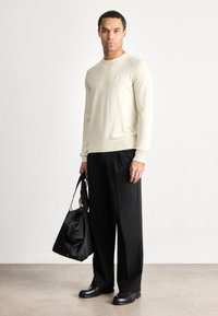 Mann trägt einen cremefarbenen Pullover und schwarze Hose, hält eine schwarze Tragetasche und steht auf einem grauen Boden vor einer weißen Wand.
