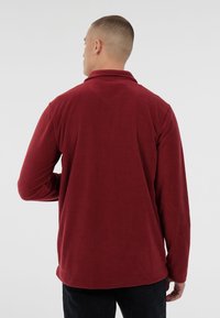 Man met kort haar, gekleed in een lange mouwen bordeauxrode fleece jas en zwarte broek, met zijn rug naar de camera gekeerd tegen een effen grijze achtergrond.