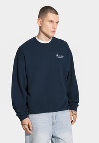 Granatowy sweter, okrągły dekolt, ściągacze przy mankietach i u dołu, haftowane logo w kolorze białym. Noszony na białej koszulce, w połączeniu z jasnymi dżinsami.