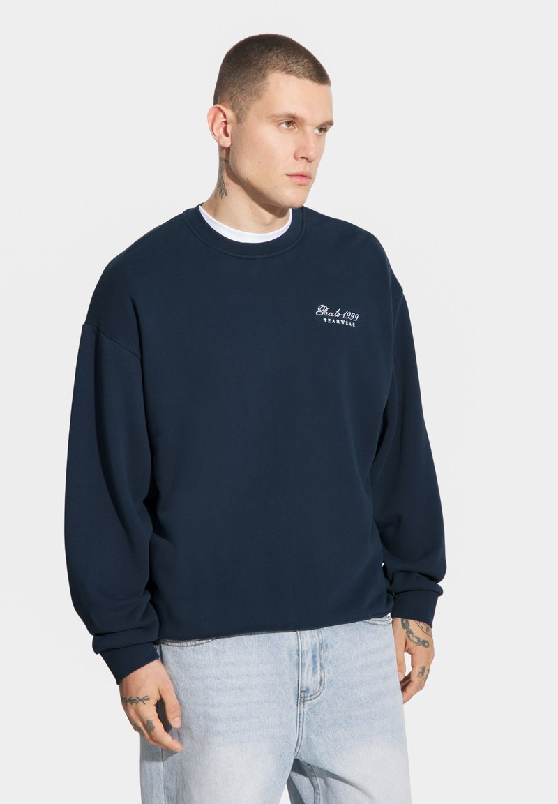 Granatowy sweter, okrągły dekolt, ściągacze przy mankietach i u dołu, haftowane logo w kolorze białym. Noszony na białej koszulce, w połączeniu z jasnymi dżinsami.